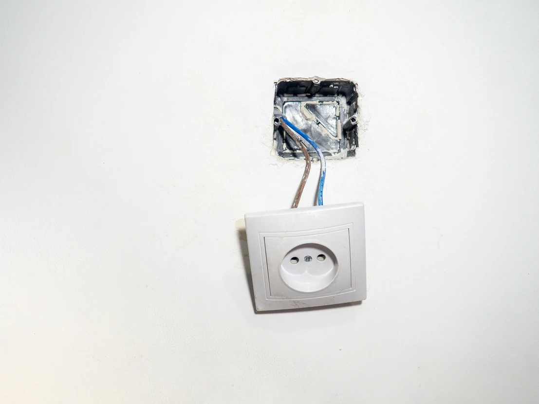 Scène d'installation électrique en intérieur : prise de courant européenne démontée avec fils électriques apparents sortant du boîtier d'encastrement mural sur fond blanc
