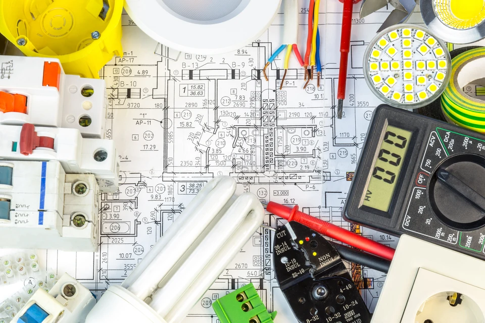 Espace de travail d'électricien avec plans techniques détaillés, multimètre numérique affichant des mesures, disjoncteurs modulaires, LED circulaire, câbles électriques colorés et composants électroniques