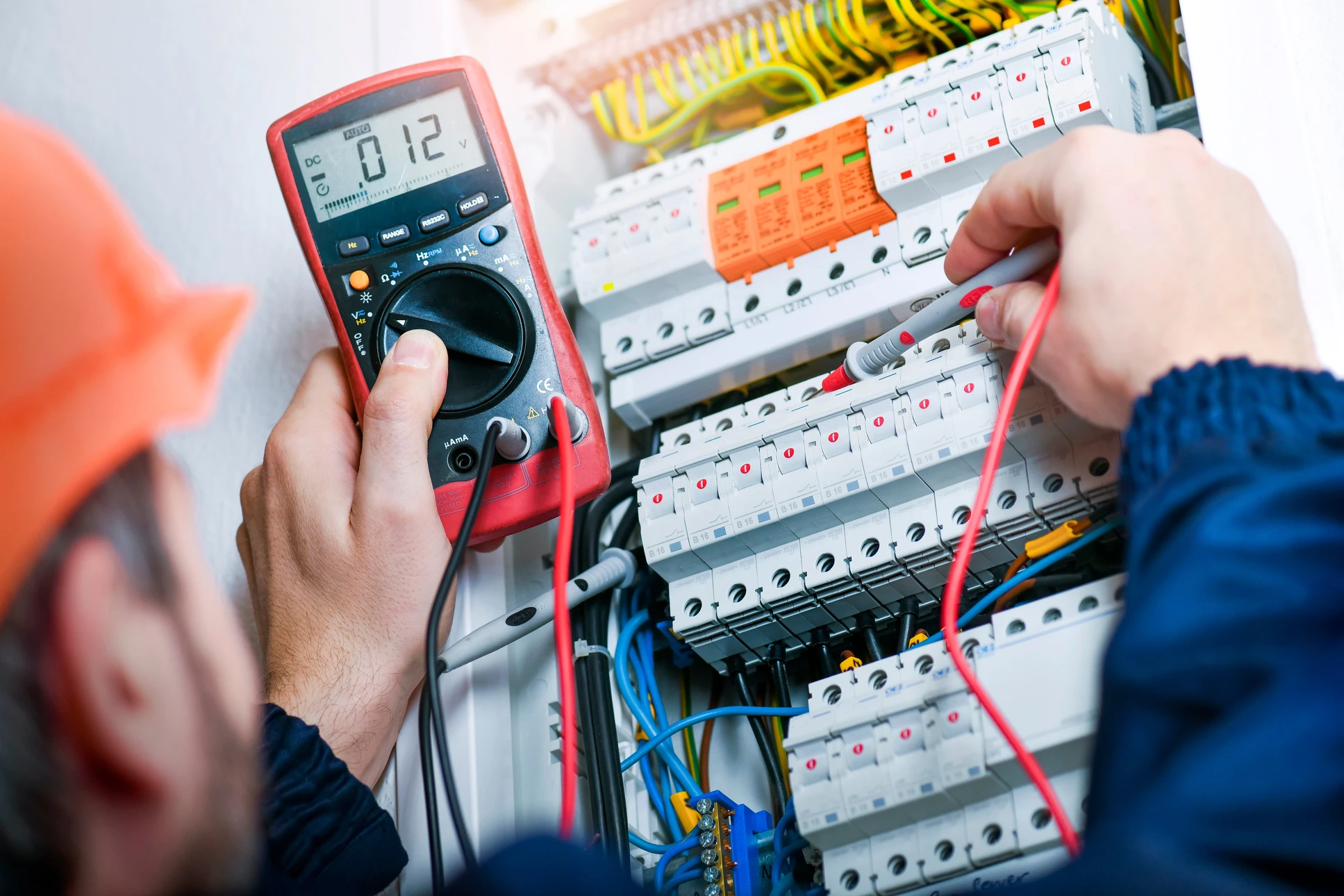Électricien en combinaison orange travaillant sur un tableau électrique résidentiel, utilisant un multimètre numérique pour effectuer des mesures de tension