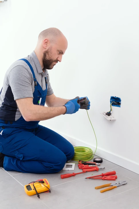 Électricien en combinaison bleue et casque orange effectuant des mesures sur un tableau électrique résidentiel